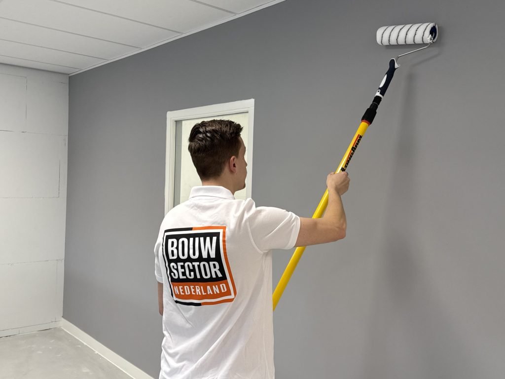 Schilder met het Bouwsector Nederland-logo rolt een grijze wand onder een systeemplafond, naast een deur met glas