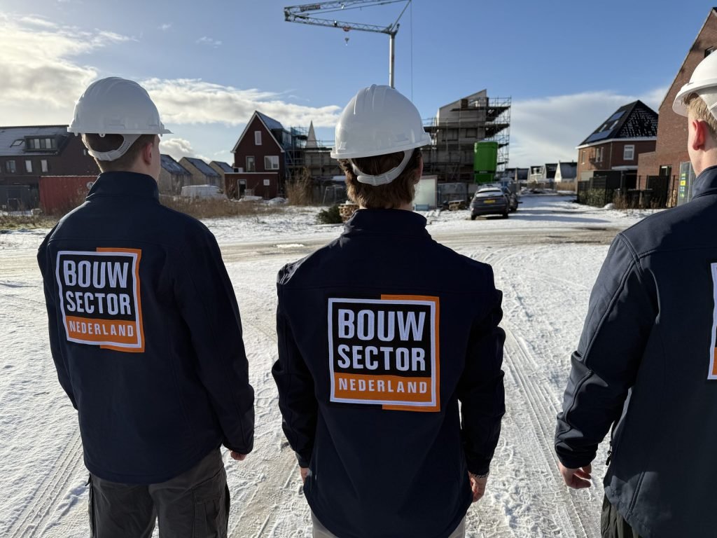Drie vakmensen van Bouwsector Nederland met witte helm kijken naar woningen in aanbouw op een besneeuwde bouwplaats