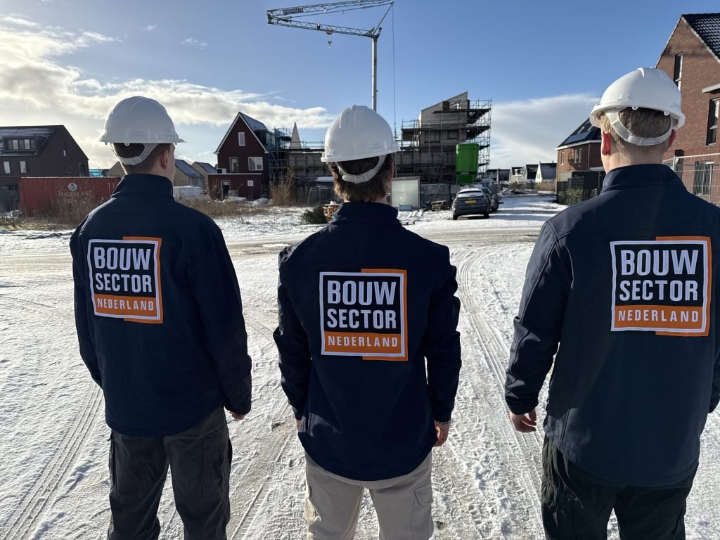 Drie vakmensen van Bouwsector Nederland met bouwhelm, op de rug gezien, op een besneeuwde straat tussen woningen in aanbouw